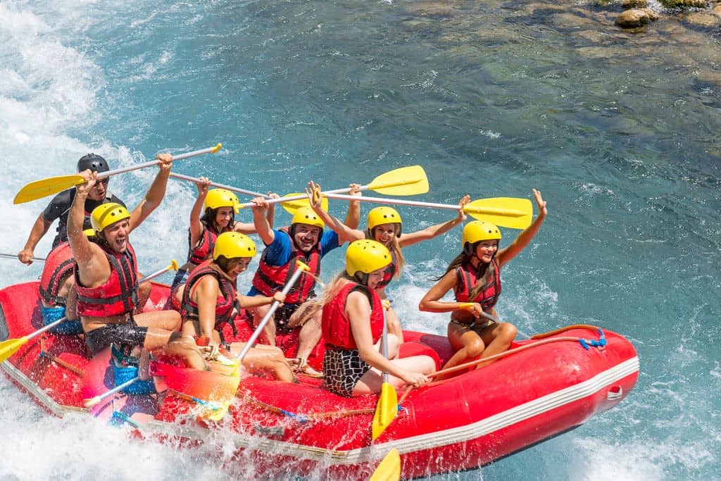 Red raft with 8 people using red life jackets and yellow paddles on a blue river