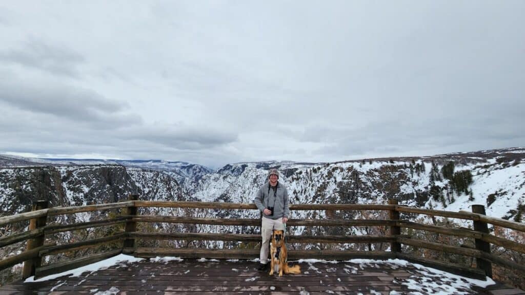 Man and dog smiling on a wooden boardwalk overlooking snowy mountains