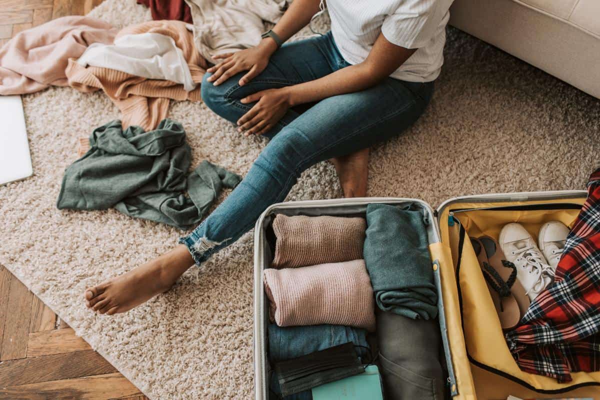 Person sitting on a carpeted floor next to a packed suitcase with clothes strewn around the floor