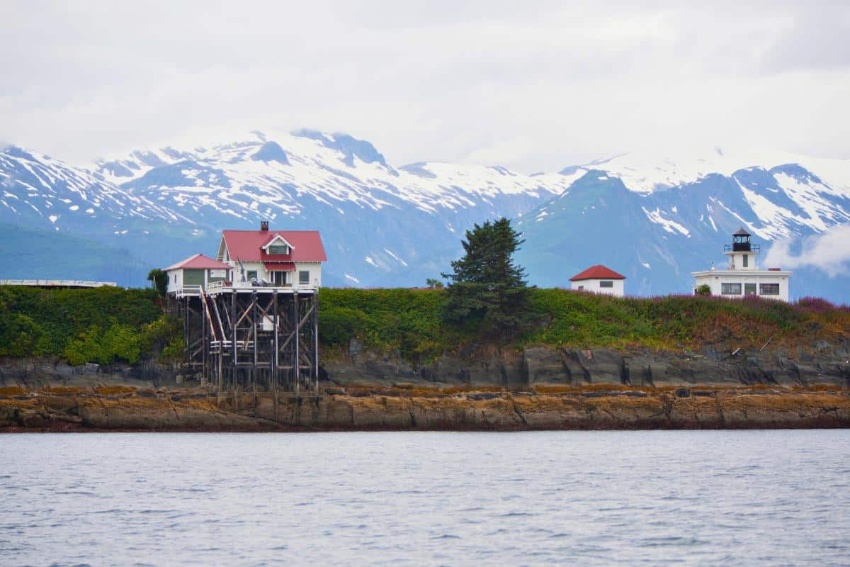 Red and white buildings with a small lighthouse on a rocky cliffside backed by snowy mountains