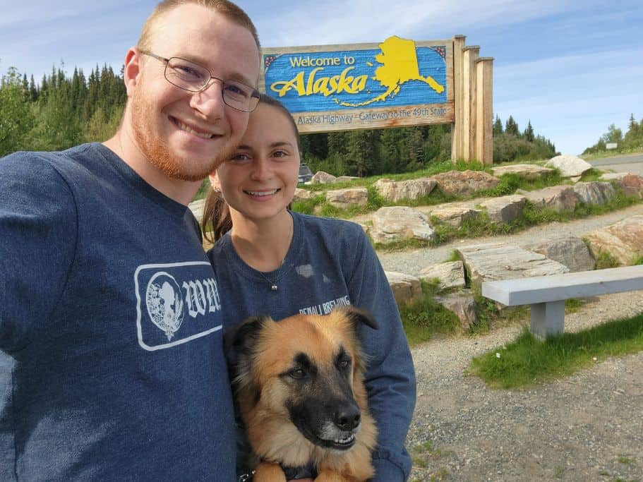 Two people and a dog smiling in front of a "Welcome to Alaska" sign