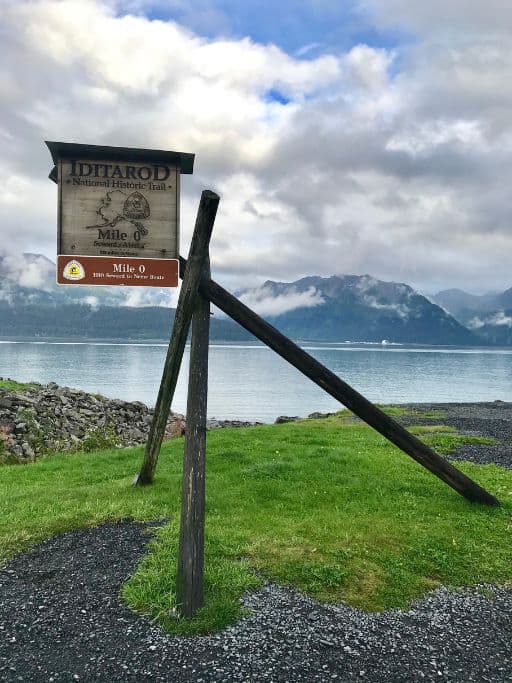 Iditarod Mile 0 sign on a grassy landing overlooking a bay backed by mountains in Alaska
