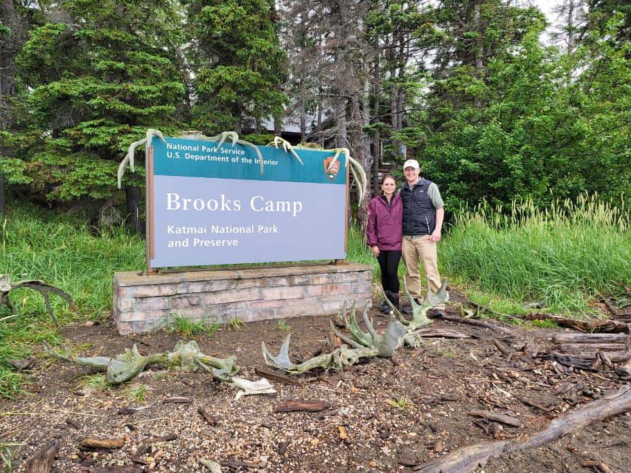 Two people standing next to a blue sign surrounded by woods that says "Brooks Camp: Katmai National Park and Preserve"