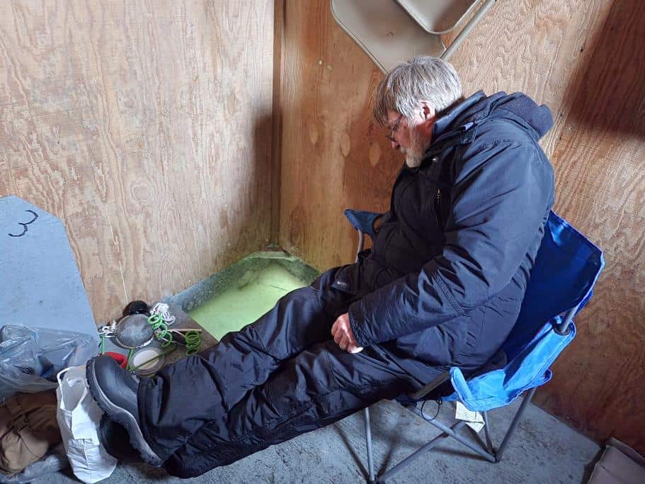 A man fishing sitting in a blue camping chair over an ice fishing hole in a cabin