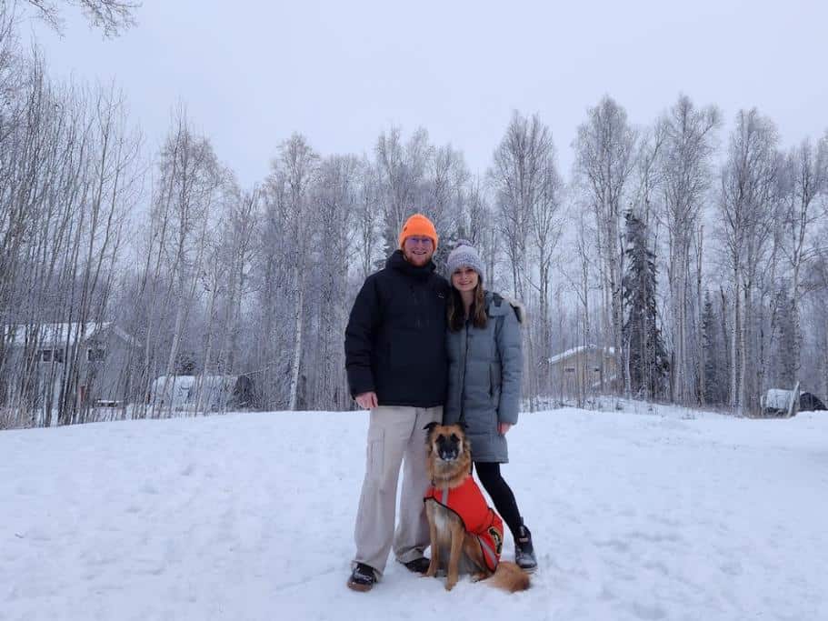 A man, woman, and dog standing in the snow surrounded by trees
