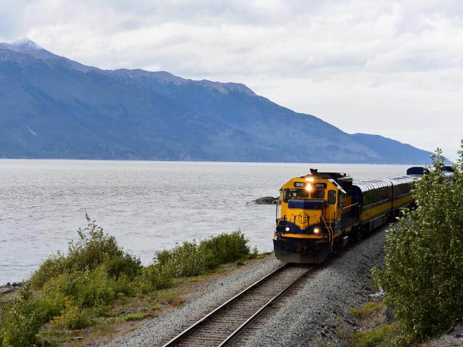 A yellow train driving along the ocean with a mountain in the background