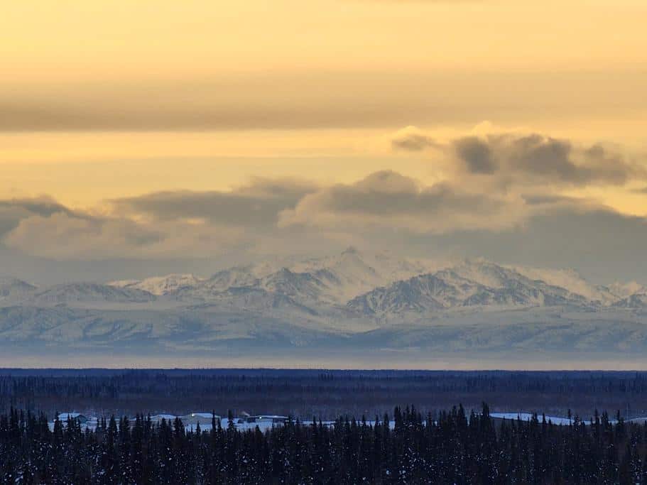 Landscape photo of snow-capped mountains, a blue lake, dark trees, and a golden sky
