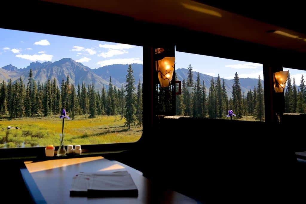 Dark interior of a train driving through mountains and pine trees with a partially sunlit table by one of the windows