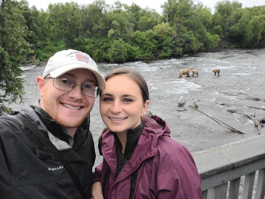 A man and woman smiling on a deck with brown bears fishing in the background