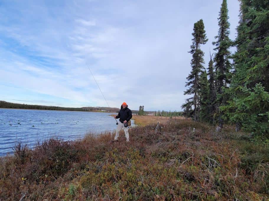 Man in a black rain coat and orange hat standing in a marsh next to a lake with a fishing rod