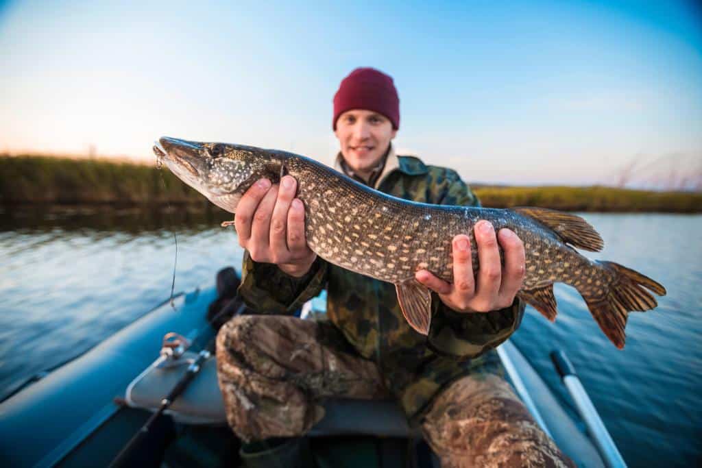 Man dressed in camo sitting in a narrow boat holding a brown spotted fish