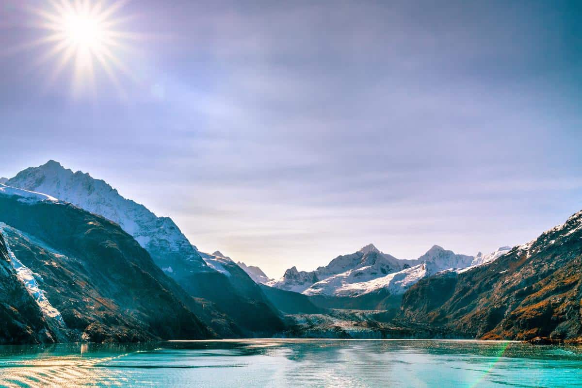 Large icy blue bay with snowy mountains and a high sun