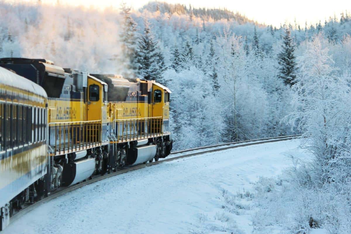 Yellow train with "Alaska" written on the side driving throw snowy terrain