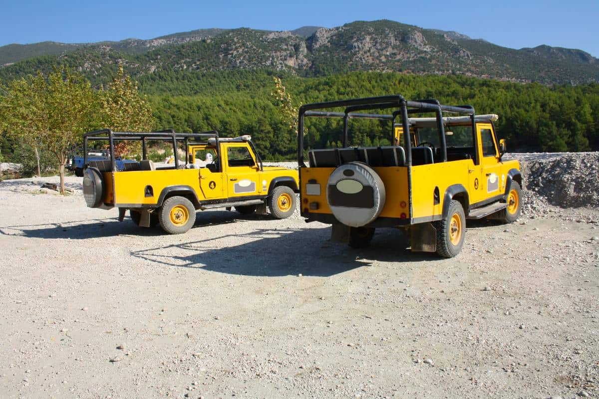 Two yellow, open-top jeeps in a dirt parking lot in front of a green mountain