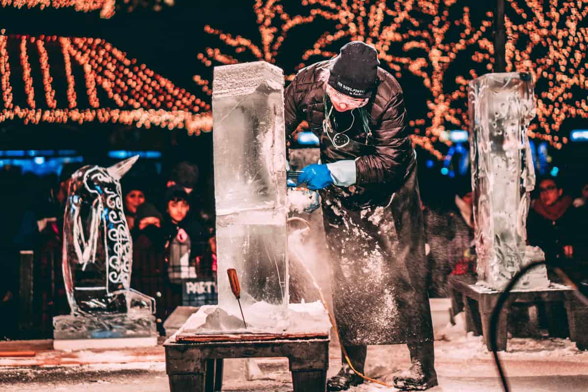 Man using a saw to shape ice under illuminated trees in the winter