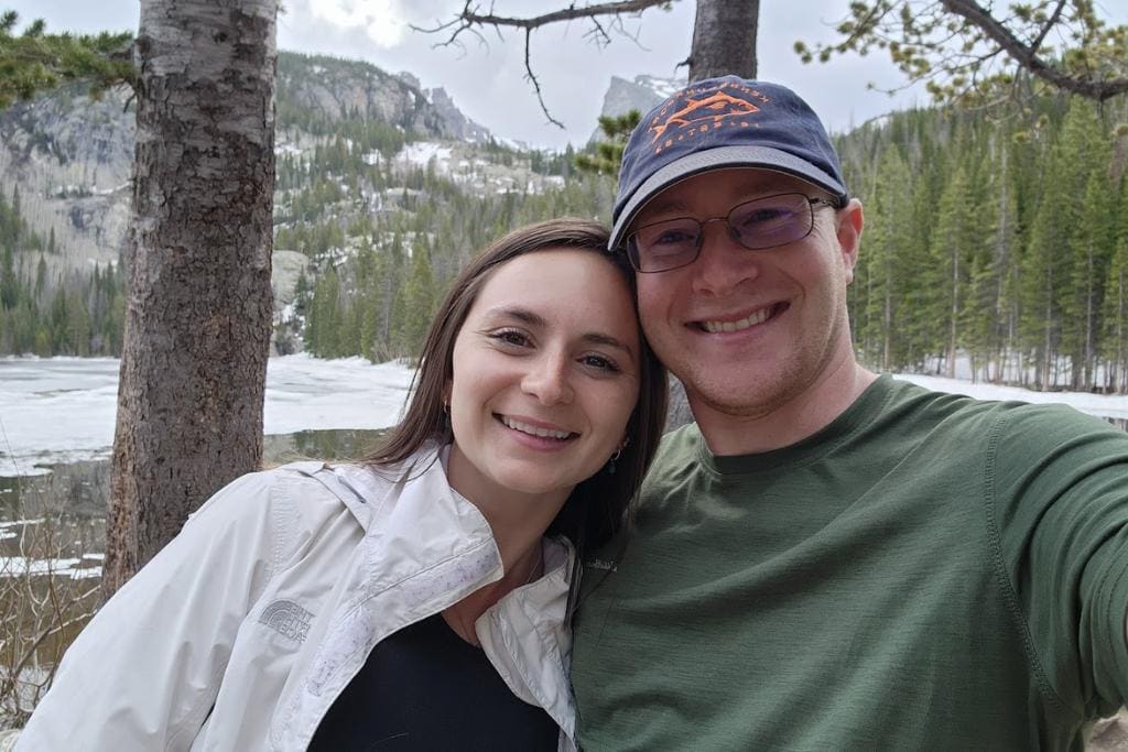 Man and woman smiling in white and green tops in front of trees and mountains