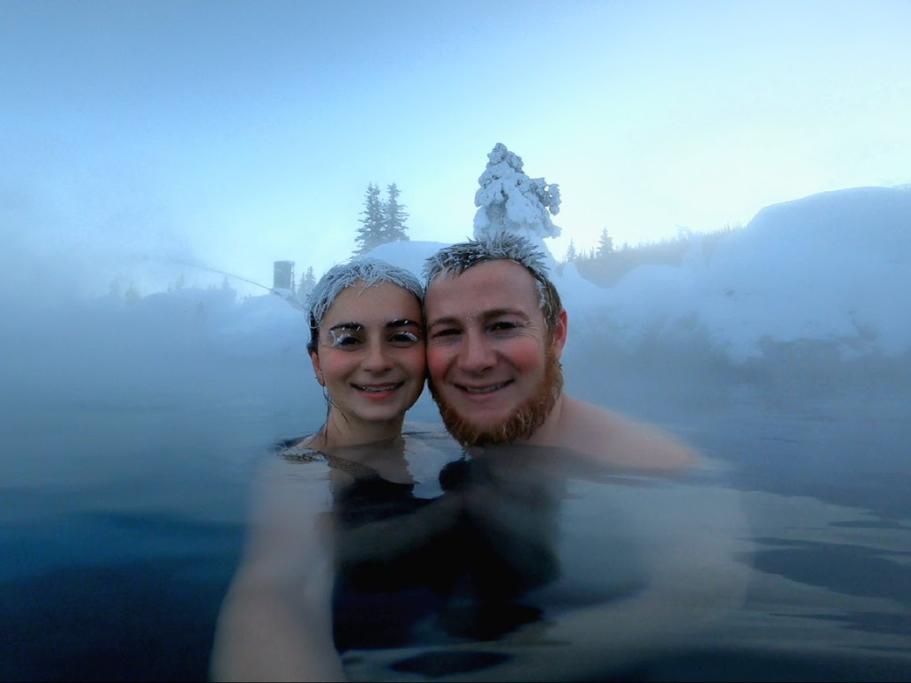 Two people in a hot spring with frosty hair surrounded by stea, and snowy trees