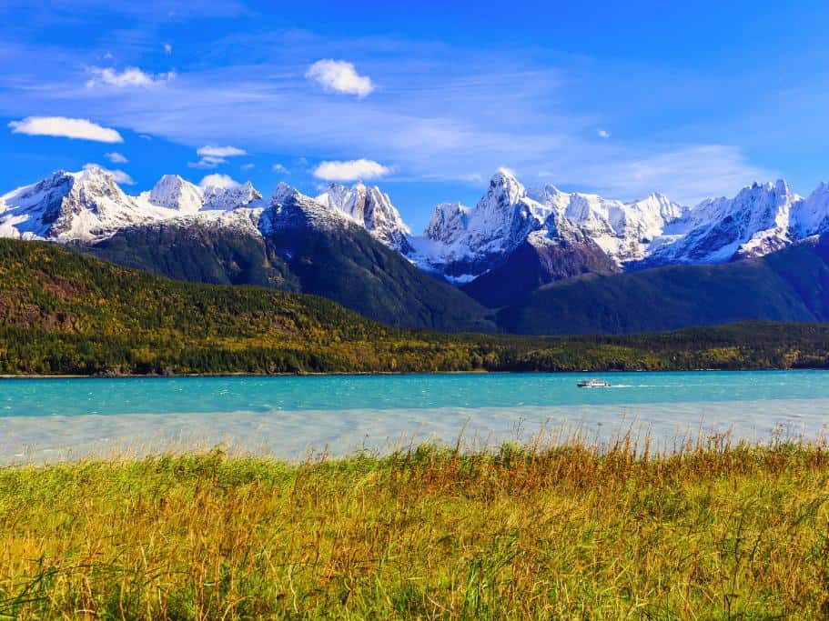 Vibrant blue water with marsh on one side and snowy mountains on the other with a boat cruising up the channel