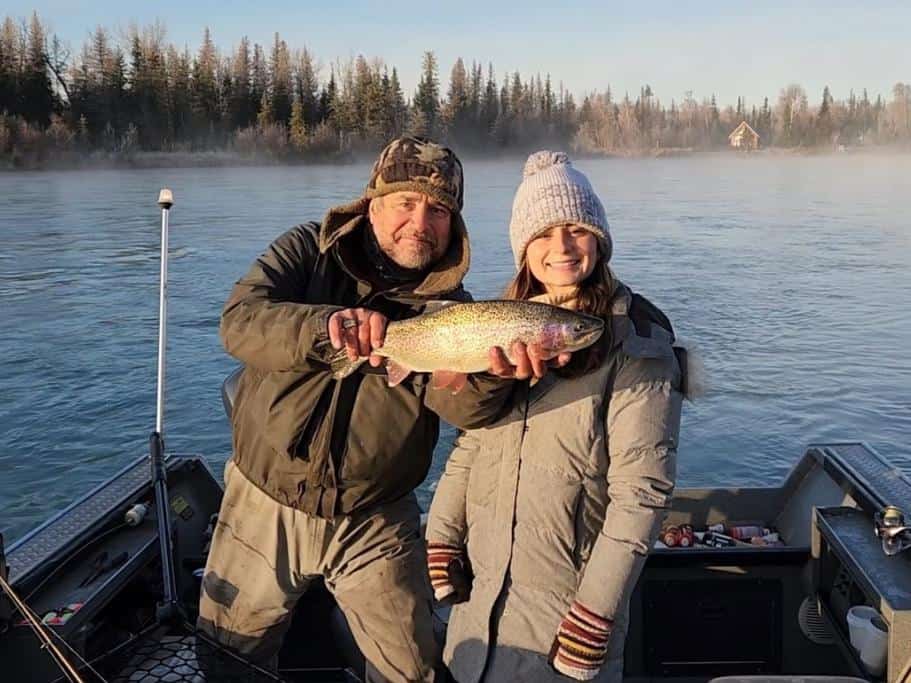 Man holding a fish next to a girl on a small boat on a wide river surrounded by pine trees and fog