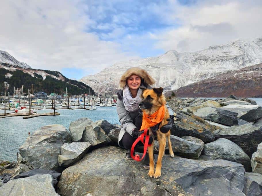 Girl and dog on a rocky jetty cutting through a small harbor backed by snowy mountains