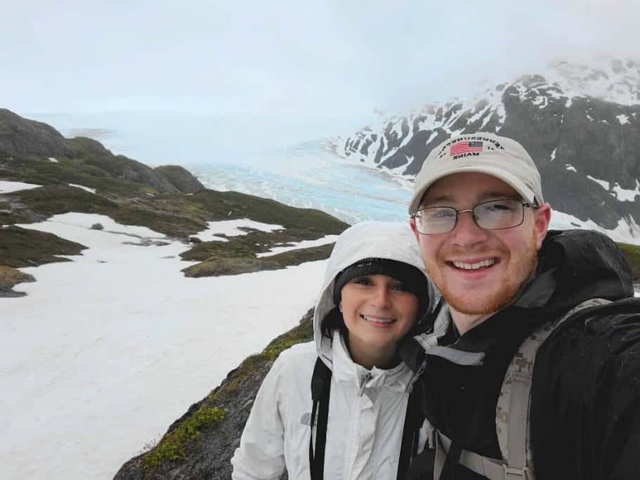 Man and woman in rain jackets smiling in front of a glacier on a snowy mountain trail