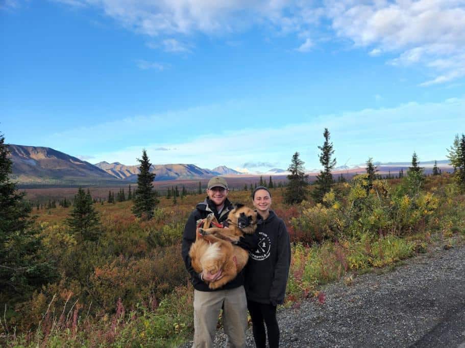 Two people holding a dog surrounded by trees and mountains
