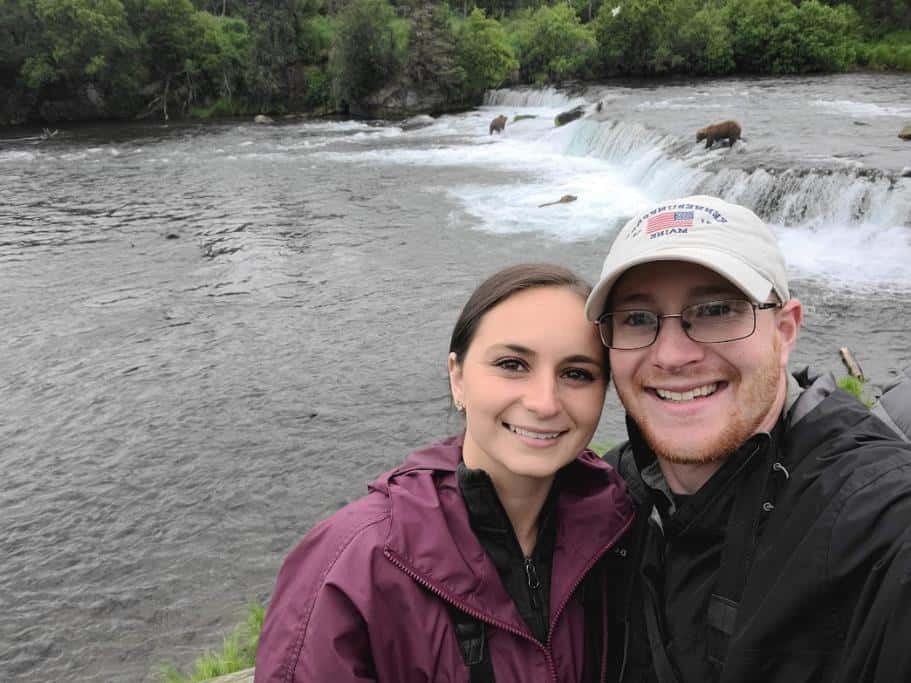 Two people in rain jackets smiling with bears fishing in a waterfall behind them