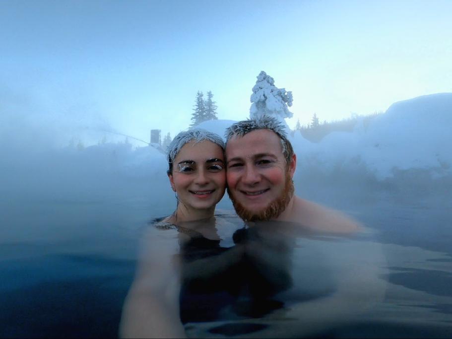 Man and woman with ice and frost in their hair while sitting in a hot spring surrounded by snowy trees and steam