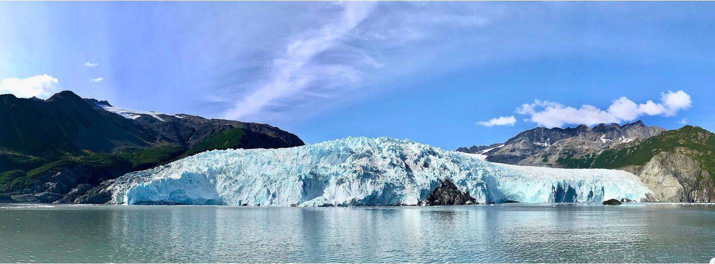 Wide angle photo of a large, blue glacier on the ocean surrounded by rocky mountains