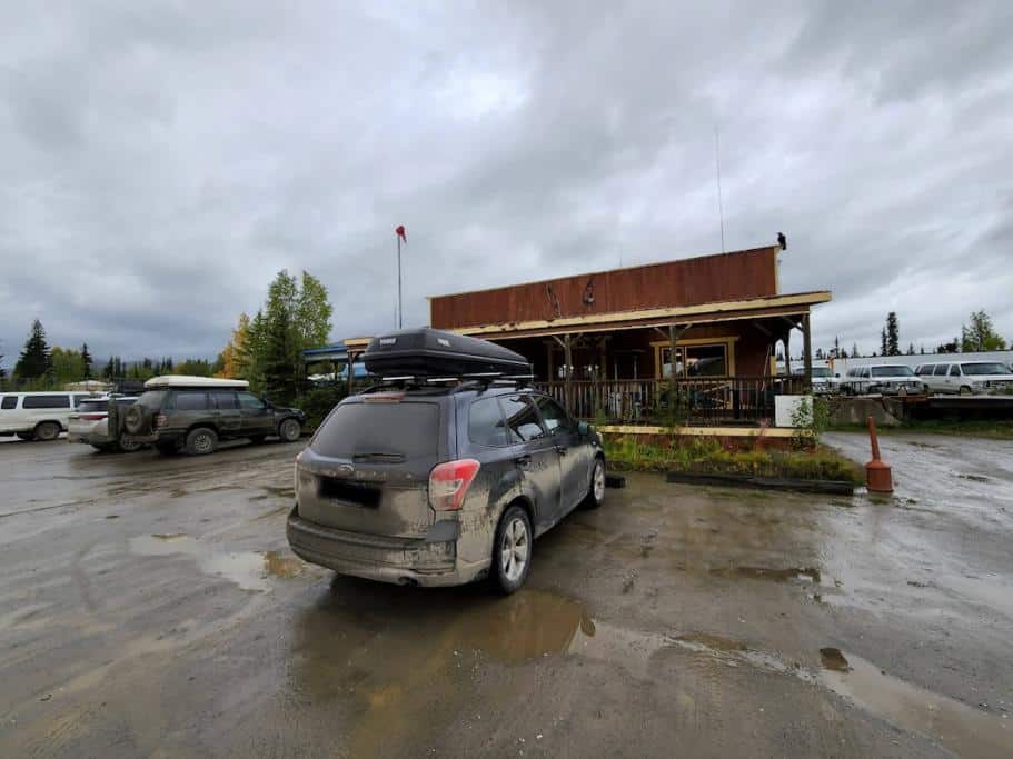 SUV parked in a muddy paring lot in front of a small, wooden building with other vehicles
