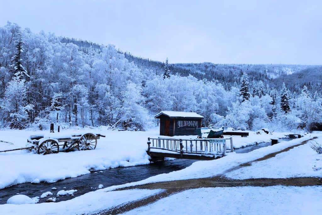Small wooden cabin with a bridge going over a dark river surrounded by snow and trees