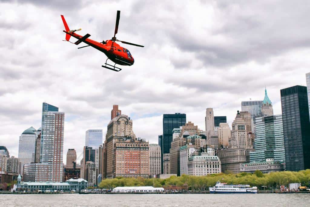 Red helicopter flying over water towards a city full of skyscrapers