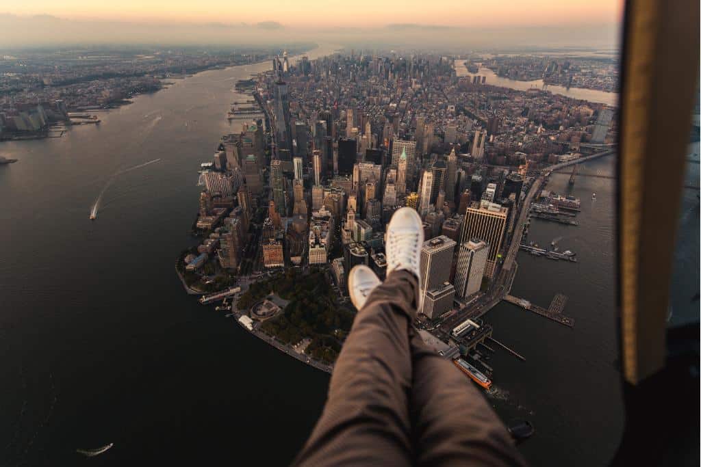 Person's legs hanging out the door of a helicopter flying over a large city surrounded by water