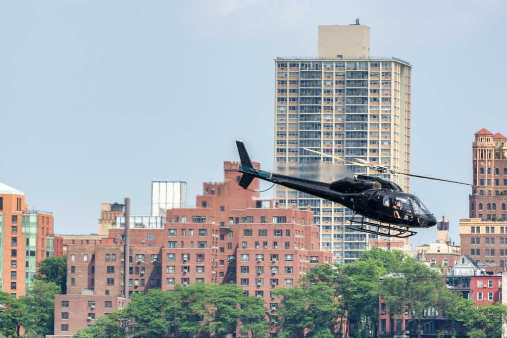 Black helicopter flying by tall brick buildings and over trees