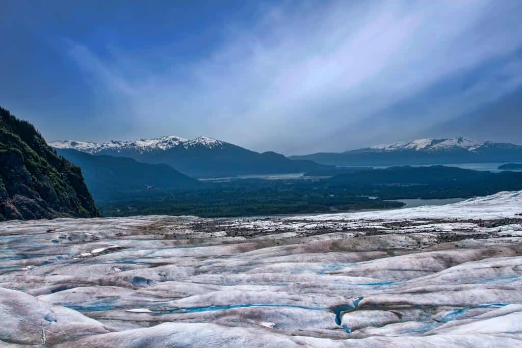 Dirty glacier top with bright blue crevasses with large mountains in the distance