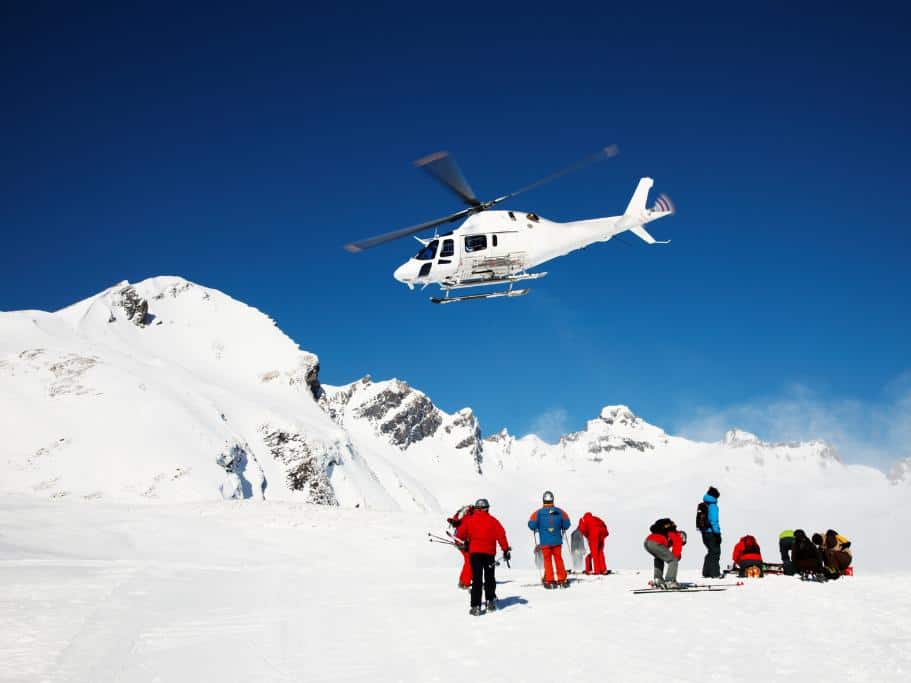 White helicopter flying over snow-covered mountains with a group of skiers in red and blue clothing