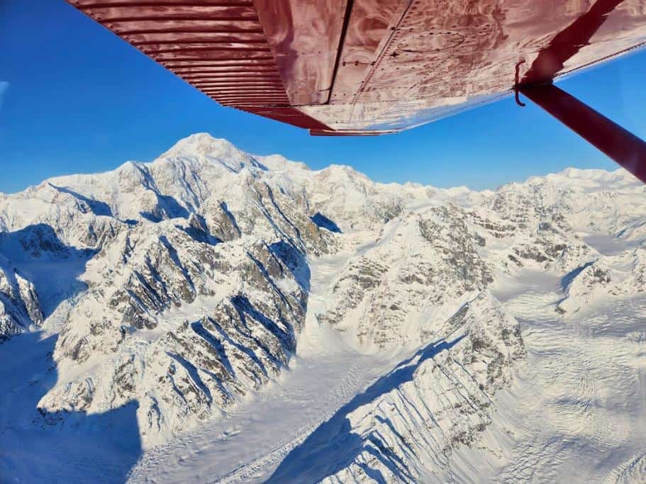 A red airplane wing with a blue sky and snowy mountains over a glacier