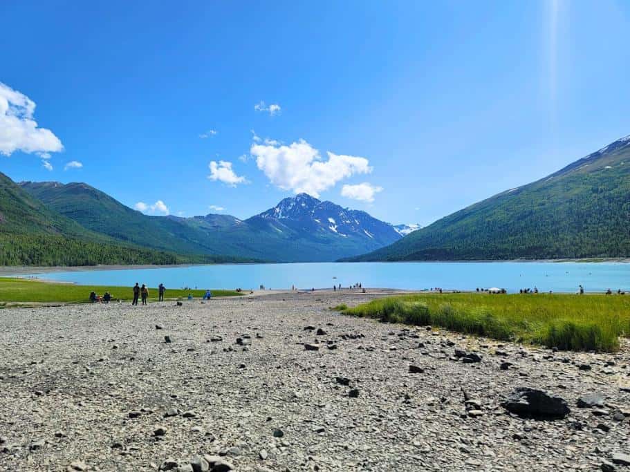 Bright blue sky with a blue lake, rocky beach, and lush mountains