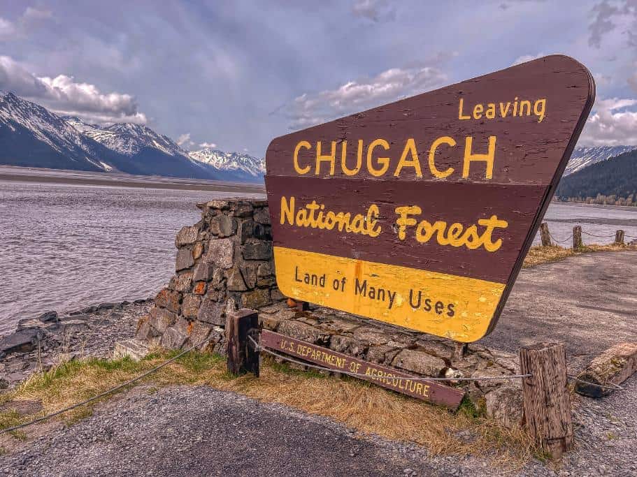 Brown and yellow "Leaving Chugach National Forest: Land of Many Uses" sign next to the ocean and mountains
