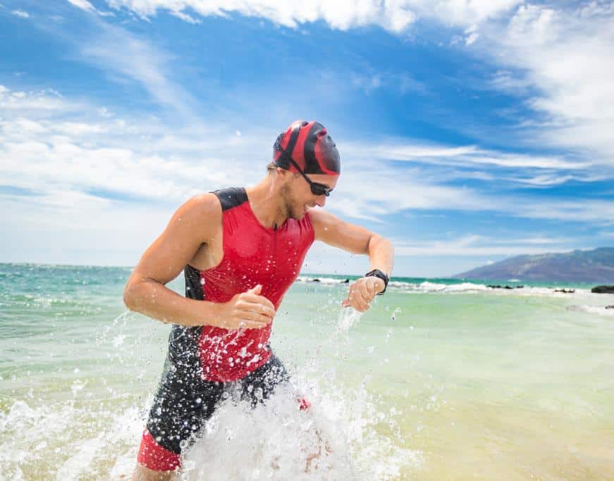 Man in a full-body swimsuit and hair cap with goggles coming out of the ocean and checking his watch