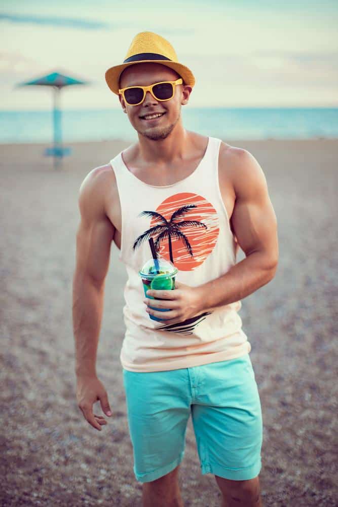 Muscular man in a tank top, blue shorts, sunglasses, and sunhat holding a drink on the beach