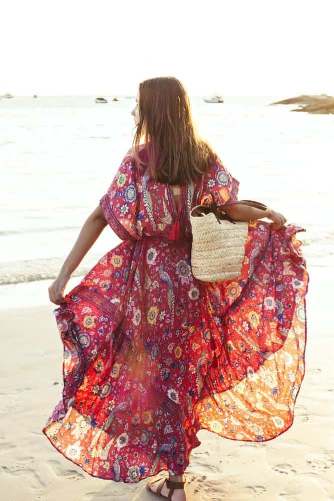 Girl in a colorful, flowy maxi dress on the beach with a beige bag