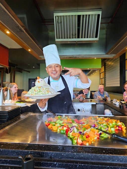 Hibachi chef next to sauteed vegetables in the shape of a heart while holding a plate of vegetable fried rice in the shape of a chicken