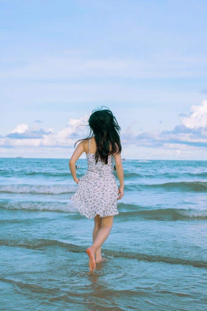 Girl with brown hair running into beach waves in a white and blue sundress
