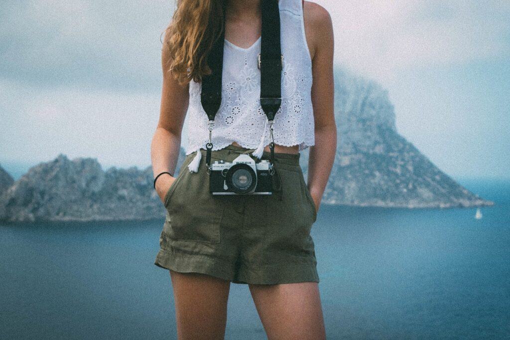Girl with a white blouse, green shorts, a camera around her neck, and mountains in the distance
