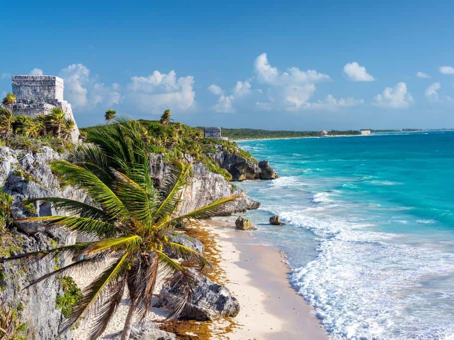Mayan ruins on a cliff next to turquoise ocean water and palm trees