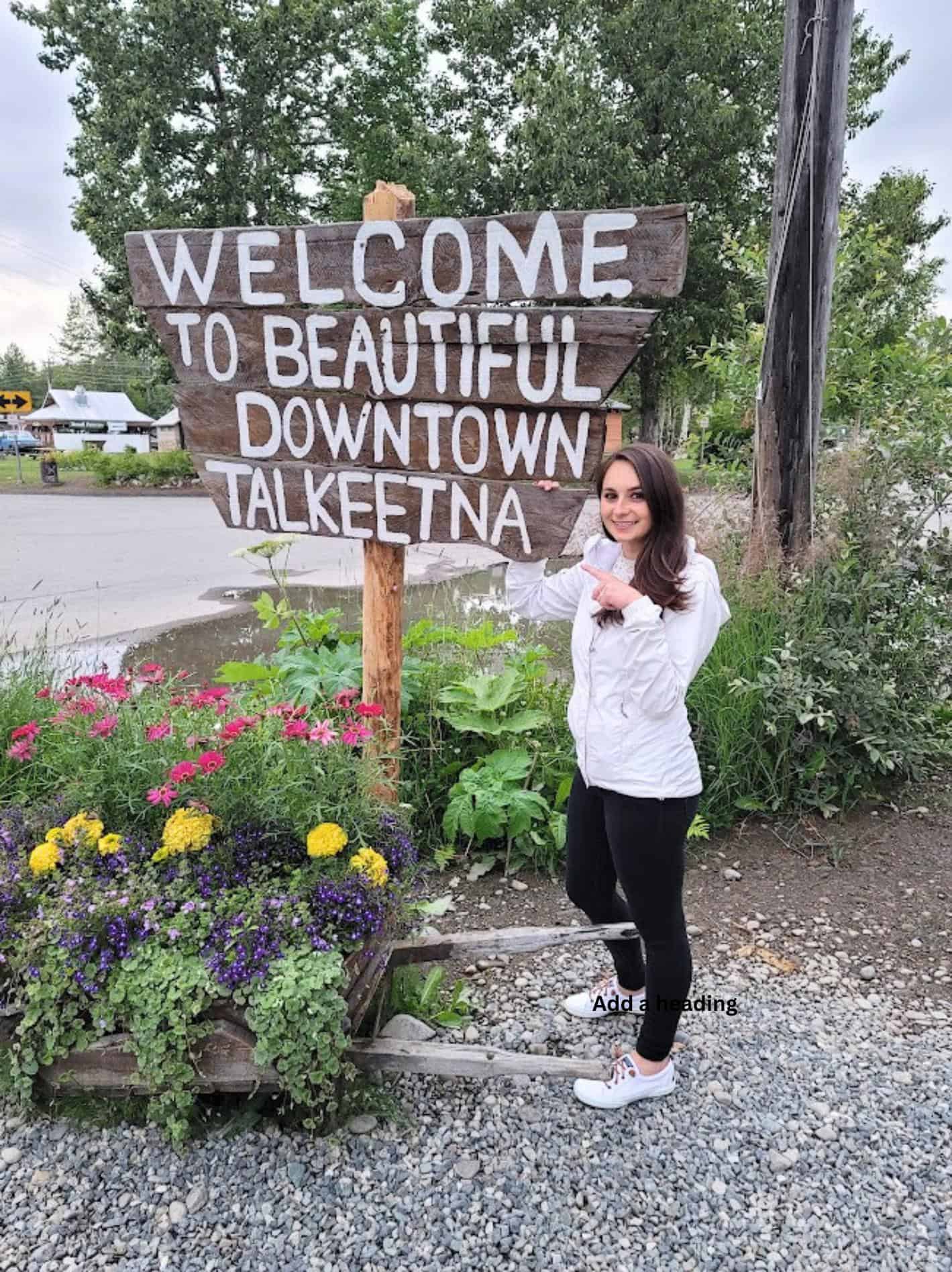 Emily Concannon next to a wooden sign saying "welcome to beautiful downtown Talkeetna"