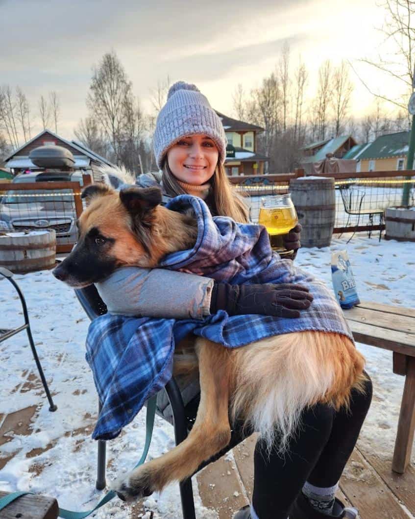 Girl with a cider and her dog on her lap on a snowy deck at Denali Brewing Co. in Talkeetna, AK