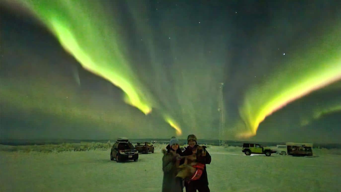 Two people holding a dog on a big mountain top with northern lights above them in Fairbanks, AK