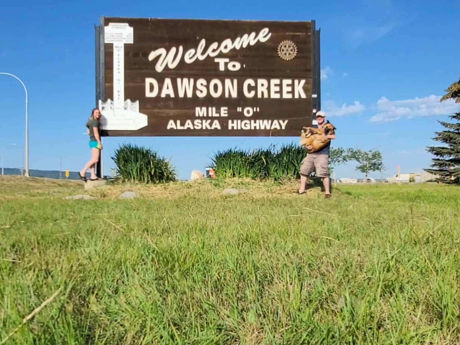 Two people and a dog in front of the "Welcome to Dawson Creek" sign in Yukon